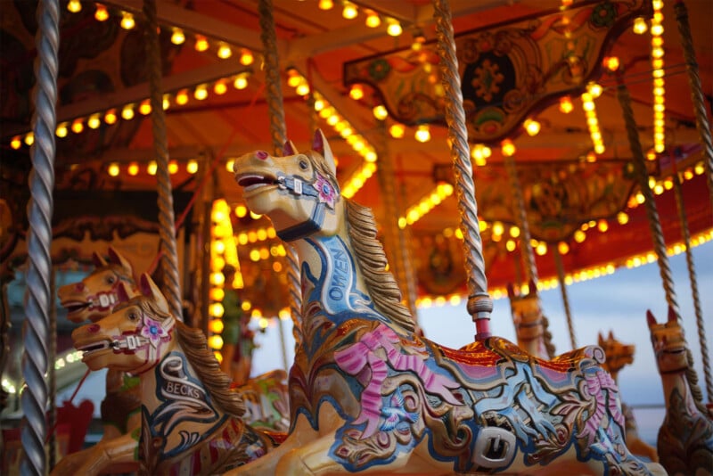 A close-up shot of carousel horses brightly lit by rows of small, warm lights. The carousel horses are intricately decorated with colorful designs, including detailed saddles and bridles. The background shows more carousel details and a glimpse of the sky.