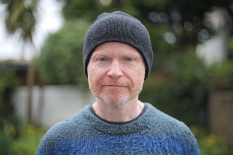 A person with a ginger beard and mustache wearing a black beanie and a textured blue sweater stands outdoors, looking directly at the camera. The background is blurred, showing greenery and a palm tree.