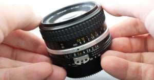 Close-up of hands holding a camera lens. The lens features various markings, including numbers representing aperture settings (from f/1.4 to f/16) and distance measurements in meters and feet. The focus ring and other detailed components are prominently visible.
