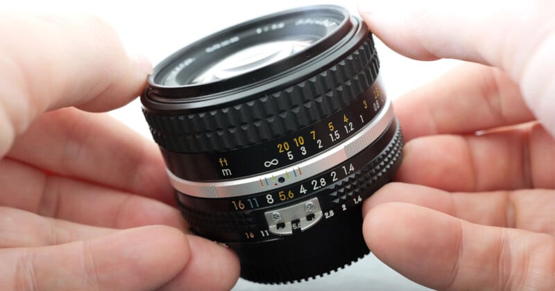 Close-up of hands holding a camera lens. The lens features various markings, including numbers representing aperture settings (from f/1.4 to f/16) and distance measurements in meters and feet. The focus ring and other detailed components are prominently visible.
