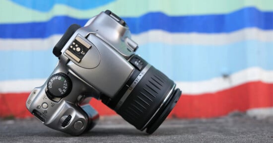 A silver DSLR camera with a black lens is laid on its side against a colorful background of horizontal stripes in blue, teal, and red. The focus dial and flash mount are visible, and the ground is slightly blurred in the foreground.