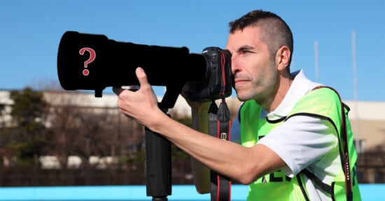 A man wearing a fluorescent green vest looks through a large camera lens mounted on a tripod. The lens has a question mark on it. The background shows a clear blue sky, trees, and buildings.