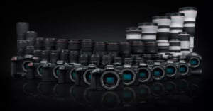 Image showing a collection of Canon cameras and lenses. The front row features several Canon camera bodies lined up side by side. Behind them, an array of Canon lenses of various sizes is displayed, gradually increasing in size from left to right. The background is black.