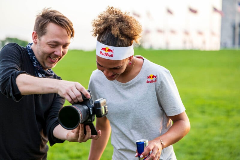 Two people standing outside on a grassy area. The person on the left shows a camera screen, sharing a moment with the person on the right, who is holding a can of Red Bull and wearing a branded headband and t-shirt. Both are smiling and appear to be having fun.