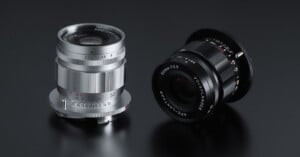 An image showing two camera lenses on a dark, reflective surface. The lens on the left is silver with white and red markings, while the lens on the right is black with white markings. Both lenses have "Voigtländer" branding visible on them.