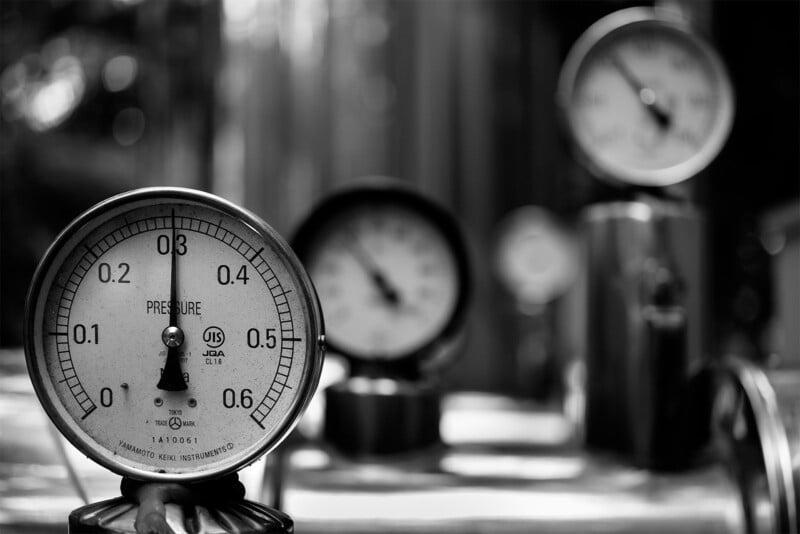 A black and white image depicting multiple industrial pressure gauges. The central gauge shows a reading of 0.4 with a clear scale. The background features two more blurred gauges, positioned at different heights, creating a sense of depth.