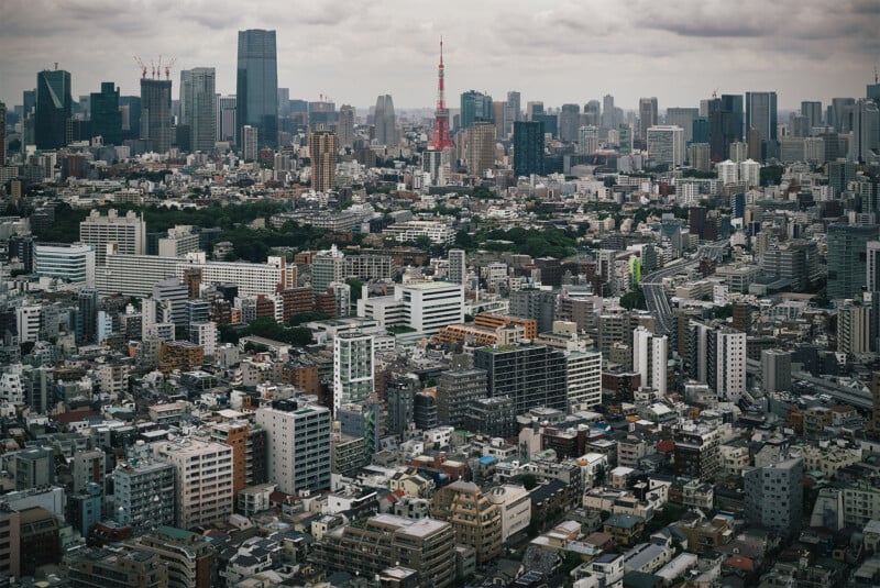 Aerial view of a dense urban cityscape featuring numerous buildings of varying heights and architectures. The iconic Tokyo Tower stands prominently in the center, surrounded by high-rise structures, under a cloudy sky.