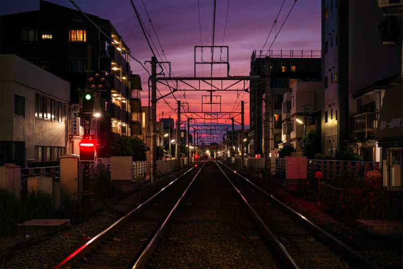 A railway track stretches into the distance at dusk, flanked by buildings on both sides. Streetlights and signal lights illuminate the scene, while the sky is painted in a gradient of pink, purple, and blue from the setting sun.