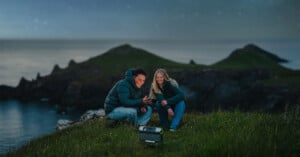 Two people are sitting on a grassy cliff at night, lit by the glow of a flashlight or a small lantern. They are looking at a device on the ground and smiling, with a calm ocean and starry sky in the background. They are both dressed warmly, suggesting a cool evening.