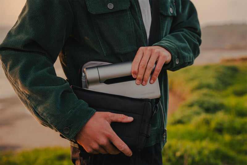 A person wearing a dark green jacket holds a compact, silver device with one hand and a black case with the other hand, standing outdoors near a grassy area, with blurred natural surroundings in the background.