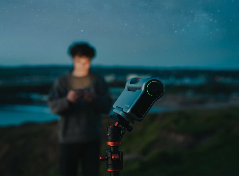 A camera mounted on a tripod is set up outdoors during twilight, with an out-of-focus person in the background. The camera lens is illuminated with a green ring, and the sky above is filled with faint stars.