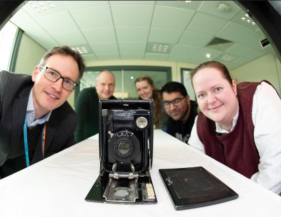 A group of five people are gathered around a table, smiling at the camera. In the foreground, there is an antique folding camera placed on the table. The background shows a modern room with large windows and fluorescent ceiling lights.