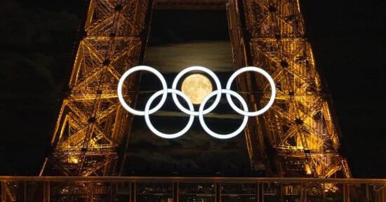 The image shows the iconic Olympic rings illuminated and suspended in front of the Eiffel Tower at night. The full moon is visible through the rings, adding a striking visual element to the scene. The Eiffel Tower's structure is lit with warm yellow lights.