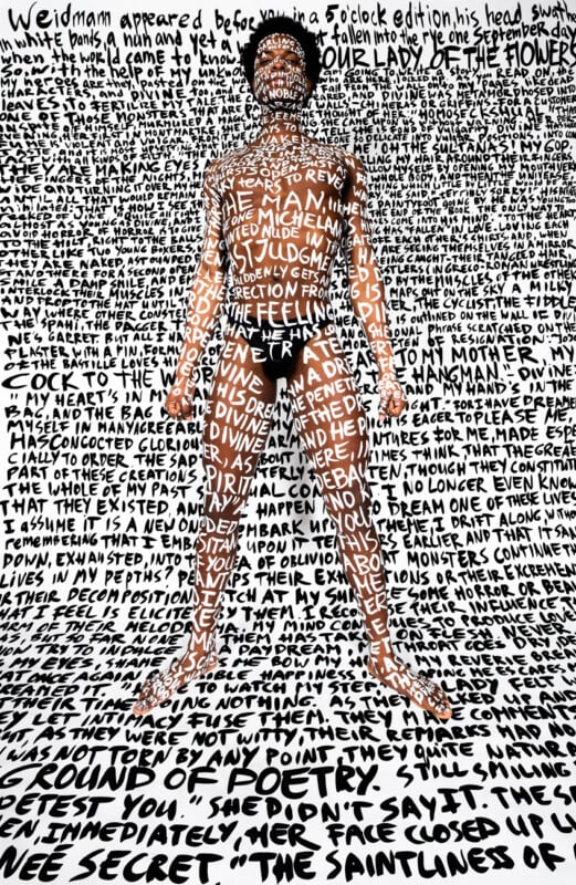 A person stands against a backdrop covered in densely packed handwritten text. The person's body is also adorned with similar handwritten text, creating a continuous, immersive effect. The text appears to cover various passages, making the figure and background blend.