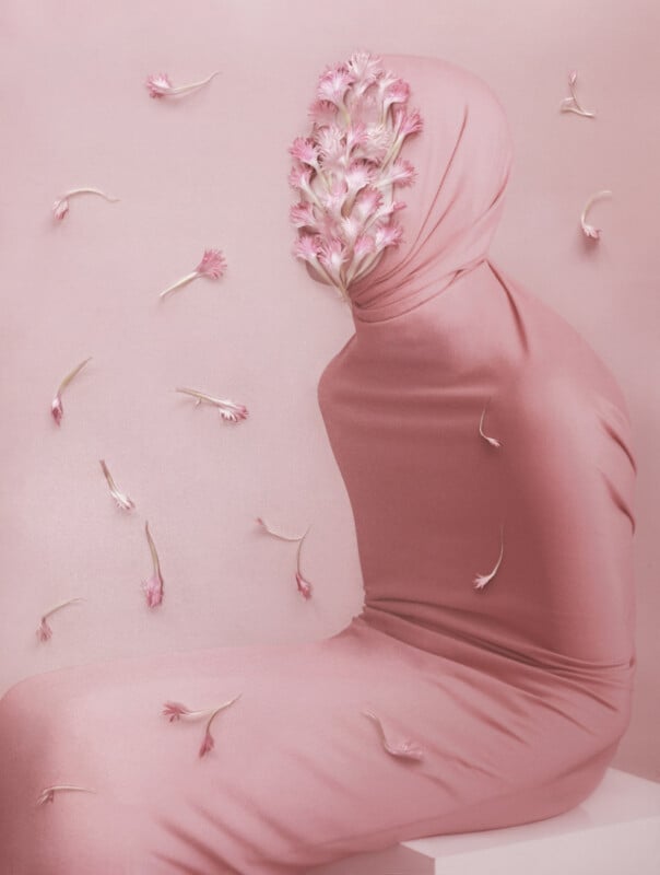 A figure covered completely in pink fabric sits on a white surface against a pink background. The face area is adorned with pink flowers. Similar flowers float around the figure. The overall aesthetic is soft and surreal.