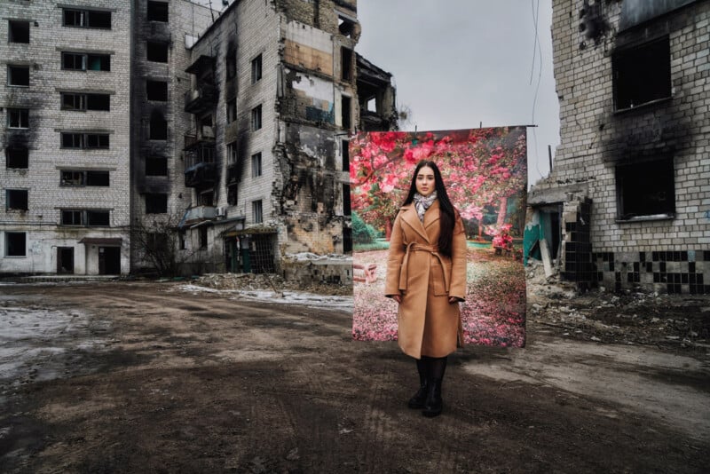 A woman stands in front of a contrast backdrop depicting a vibrant, colorful garden, which is juxtaposed against the stark, dilapidated buildings of a war-torn area. She is wearing a long, brown coat, dark pants, and black boots. The ground is barren and uneven.