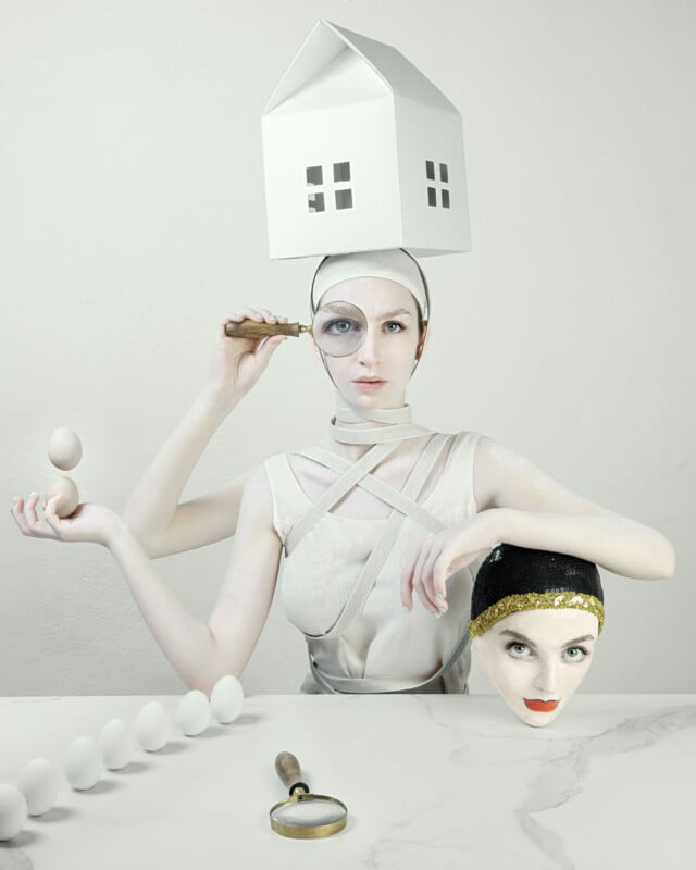 A surreal scene features a woman with a small model house on her head, holding an egg in one hand and a magnifying glass in the other. Her other hand rests on a lifelike mask of her own face. Several eggs and a magnifying glass lie on a table in front of her.