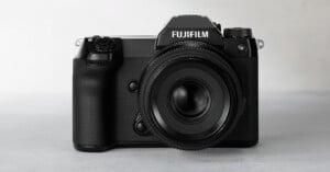 A Fujifilm digital camera with a large lens is displayed against a light gray background. The camera has a textured grip on one side and several control dials on top, with the Fujifilm logo prominently displayed above the lens.