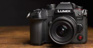 A black Panasonic LUMIX GH7 camera with a wide-angle lens is placed on a wooden surface, against a dark background. The camera has a textured grip, various buttons, and a large viewfinder. The brand name "LUMIX" is clearly visible above the lens.