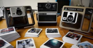 A collection of three vintage Kodak instant cameras are displayed on a wooden table surrounded by piles of Polaroid photos. The camera models include the EK4, The Handle, and the Pleaser II. The photos depict various scenes and are arranged neatly in front.