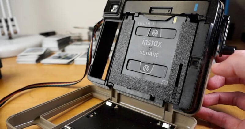 A close-up image shows the inside of an open Instax Square instant camera with a film cartridge installed. The camera is positioned on a wooden surface with photo prints and a white object blurred in the background. A hand is holding the side of the camera.