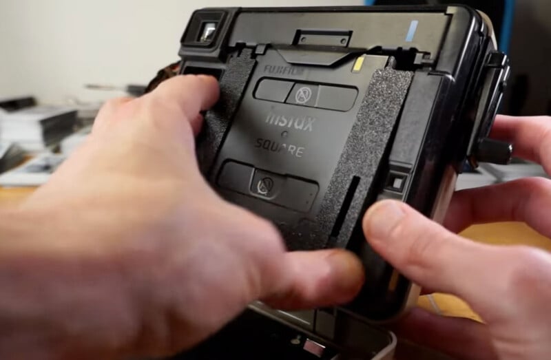 Hands are loading a film cartridge into a Fujifilm Instax Square instant camera. The cartridge is labeled with the camera model name "Instax Square." A work surface with various items is visible in the blurred background.