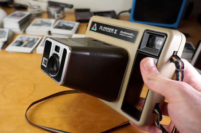 A person holding a vintage Kodak Pleaser II instant camera. The camera has a beige body with a black section around the lens and viewfinder. A colorful neck strap is attached to the camera. Polaroid photos and another camera can be seen on the table in the background.