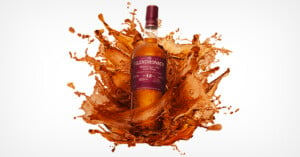 A bottle of Glendronach 12-Year-Old Single Malt Scotch Whisky with a maroon label and gold accents is shown at the center of an explosion of amber liquid splashes, set against a white background.