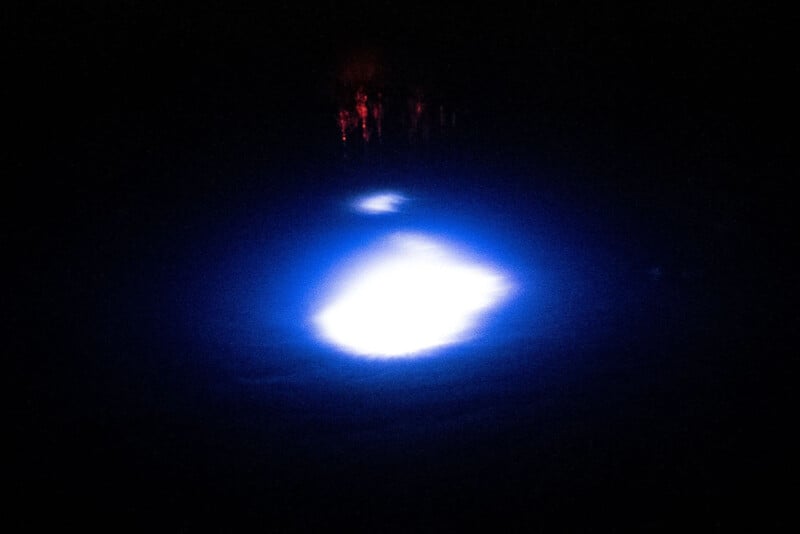 A dark scene featuring a glowing blue and white light in the center, creating an ethereal effect in the surrounding darkness. The origin of the light is not visible, but it illuminates a portion of the water surface beneath it.