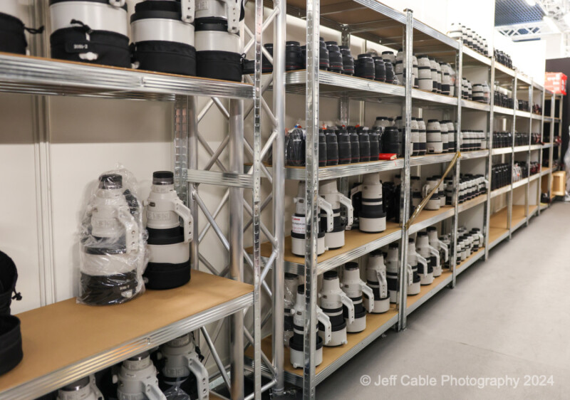 A well-organized storage area with multiple shelves holding numerous, large white camera lenses. The shelves are made of metal and the floor is smooth concrete. Some lenses have protective covers on them. The image is credited to Jeff Cable Photography 2024.