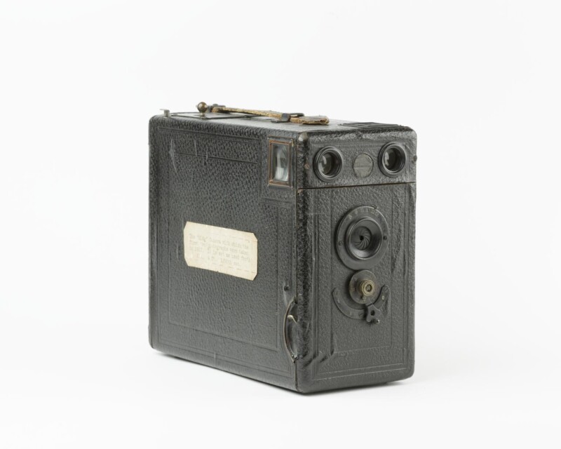 A vintage box camera with a textured black body, two circular lens openings on the front, and a small label attached to the side. The camera features a simple design with various knobs and mechanisms, indicative of early 20th-century photography equipment.