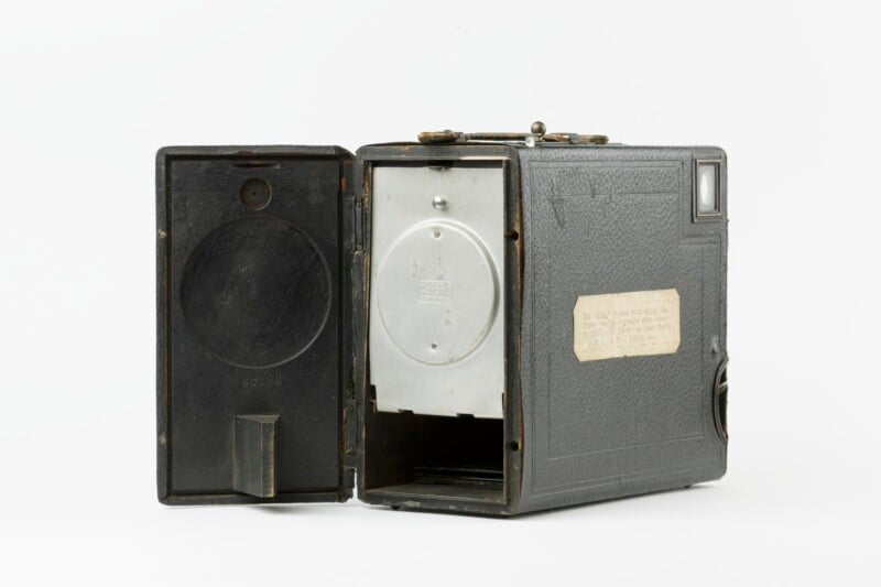 A vintage box camera with an open door revealing its interior mechanics. The exterior is textured and features a small rectangular viewfinder. A worn label is affixed to the side of the camera, and the overall color is dark, with a metallic mechanism visible inside.