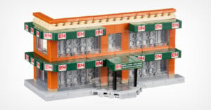 A Lego model of a two-story building with green B&H signage on multiple sides. The building features orange walls, large windows, and a small overhang above the entrance. The base is gray, mimicking a sidewalk.