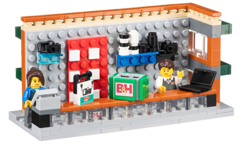 A LEGO scene depicting a camera store. A female LEGO figure is at a cash register on the left, with a male figure at a counter holding a camera. Shelves behind them display cameras, lenses, and a "B&H" logo. A laptop is on the right counter, and a window is on the right wall.