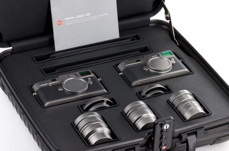 A custom-made briefcase with foam inserts containing two silver Leica cameras and three interchangeable lenses. The case includes a silver Leica 100th edition card and is secured with a combination lock.