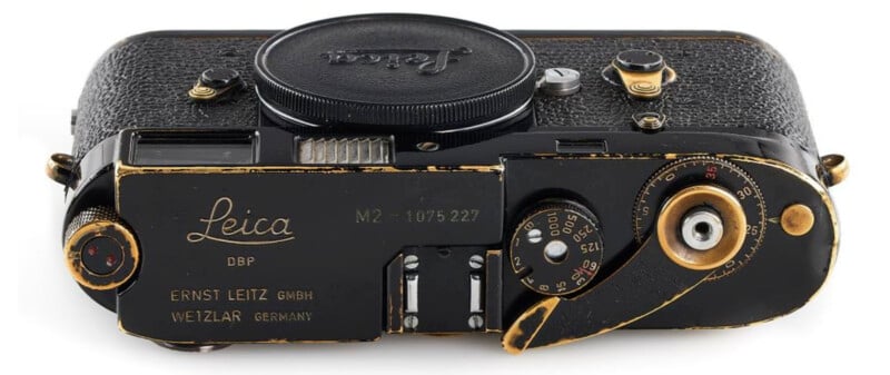 A vintage black Leica M2 film camera with visible wear and brass showing through the black paint. The top of the camera features various dials and controls. The Leica logo, model details, and manufacturing information are prominently engraved on the camera body.