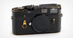 A vintage Leica film camera with a black body and brass accents, featuring a textured grip and a lens mount cap. The camera shows signs of wear, indicating frequent use. It has various buttons and dials on top and on the front, and a viewfinder on the upper right.