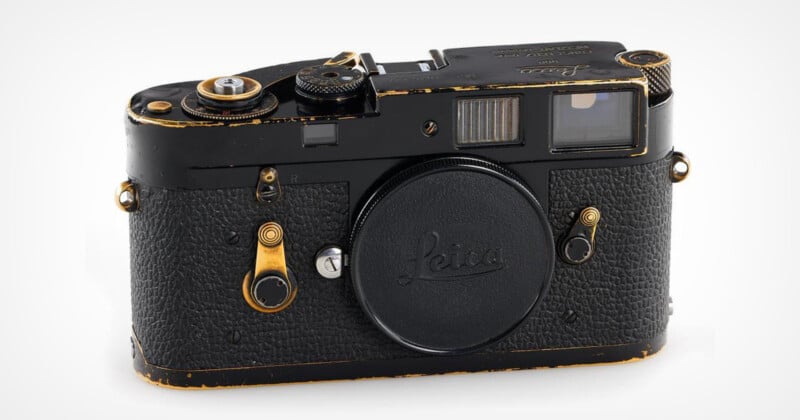 A vintage Leica film camera with a black body and brass accents, featuring a textured grip and a lens mount cap. The camera shows signs of wear, indicating frequent use. It has various buttons and dials on top and on the front, and a viewfinder on the upper right.