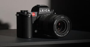 A black Leica camera with a 24-90mm zoom lens sits on a dark surface. The camera is partially illuminated from the left, emphasizing its textured grip and the iconic red Leica logo near the viewfinder.