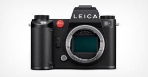 A black Leica mirrorless digital camera without a lens attached is shown. The brand name "LEICA" is prominently displayed at the top, and the camera's sensor is visible in the central mount area. The camera has a textured grip on the left side.