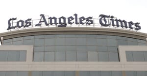 Exterior view of a building with a large sign on top that reads "Los Angeles Times" in bold, black letters. The sign sits atop a modern structure with a row of reflective windows. The overcast sky forms the backdrop.