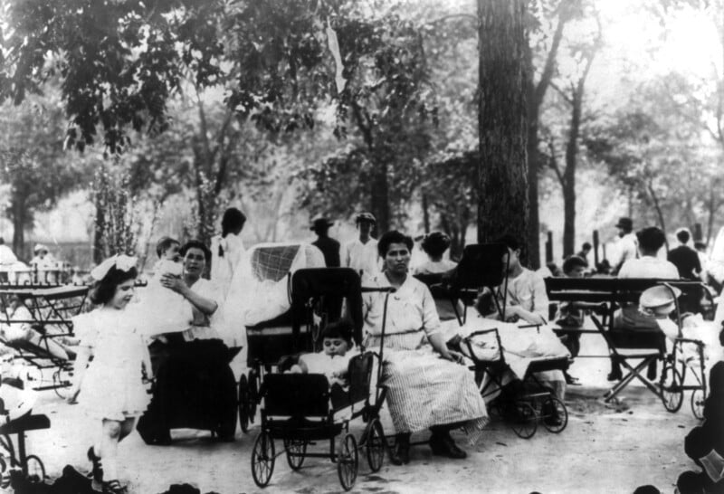 A black-and-white image of a park with several people gathered. Women are sitting on benches, some tending to children in strollers. A little girl with a large bow in her hair walks towards the camera. Trees and other park-goers can be seen in the background.