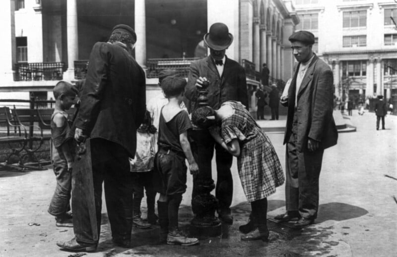 A group of children and adults gather around a public drinking fountain on a city street in the early 20th century. The children drink from the fountain while adults look on. The scene includes buildings with columns and people walking in the background.