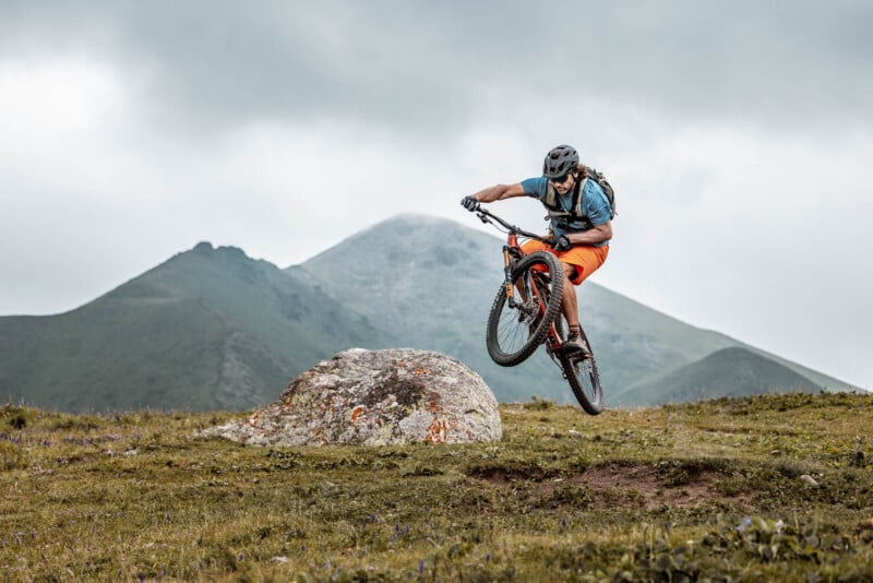 A mountain biker wearing a helmet, blue shirt, and orange shorts performs a jump over a rocky terrain with a grassy field below. In the background, there are green, rolling hills and a cloudy sky.