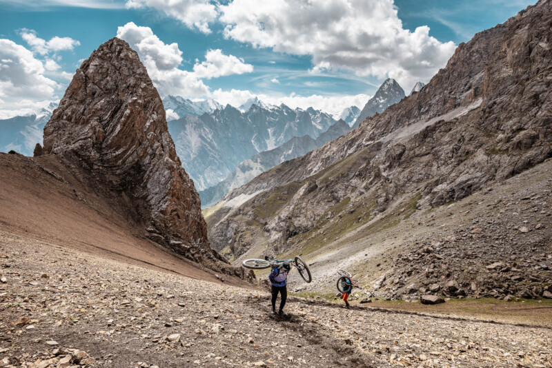 Two hikers carrying bicycles trek up a steep rocky mountainside. Towering peaks are visible in the background under a partly cloudy sky. The rugged terrain and distant snow-capped mountains create a dramatic and adventurous scene.