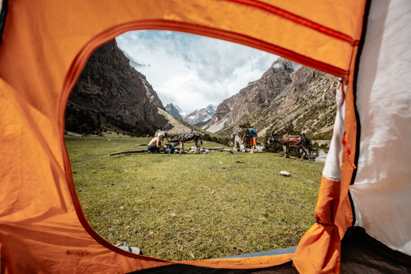 View from inside a bright orange tent, overlooking a green meadow surrounded by steep mountain ranges. People are seen in the distance with horses or mules, seemingly preparing for a trek. The sky is partly cloudy, creating a picturesque outdoor scene.