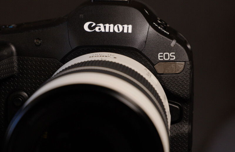 Close-up of a black Canon EOS camera with part of a white lens attached. The word "Canon" is prominently displayed on the top of the camera body, and the model designation "EOS" is visible on the right side. The background is dark and out of focus.