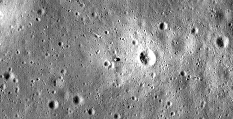 A grayscale image showing the surface of the moon with numerous craters of varying sizes. In the center of the image, there is a noticeable shadow, creating a dark spot among the surrounding terrain. The surface appears rugged and uneven.