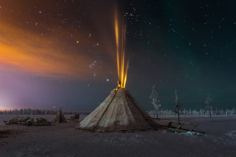 A lone tepee stands in a snowy expanse under a star-filled night sky. Warm light and smoke emanate from the top, illuminating the surroundings. The sky displays an array of colors, from deep blues and purples to subtle greens and oranges near the horizon.