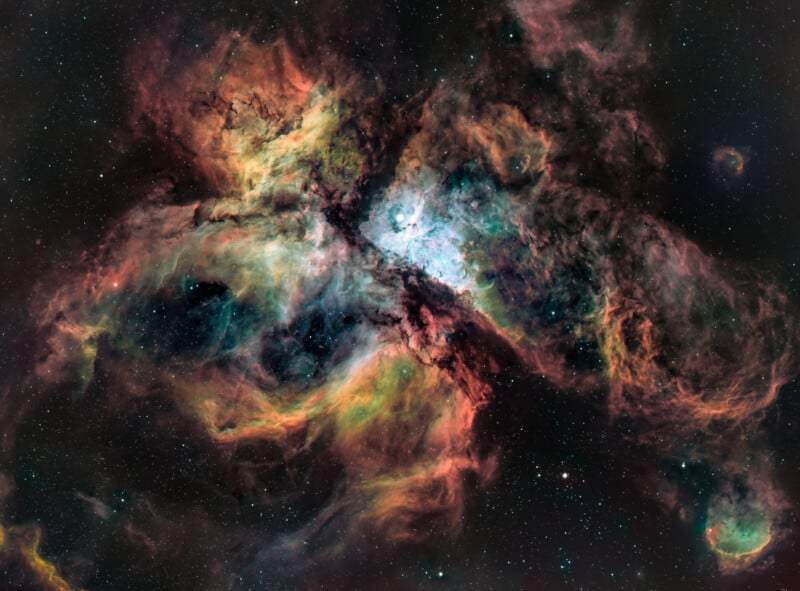 A vivid astronomical image shows the Carina Nebula in deep space. Swirling clouds of gas and dust in vibrant colors of red, orange, green, blue, and black are illuminated by nearby stars. Tiny white stars are scattered across the dark background of the universe.